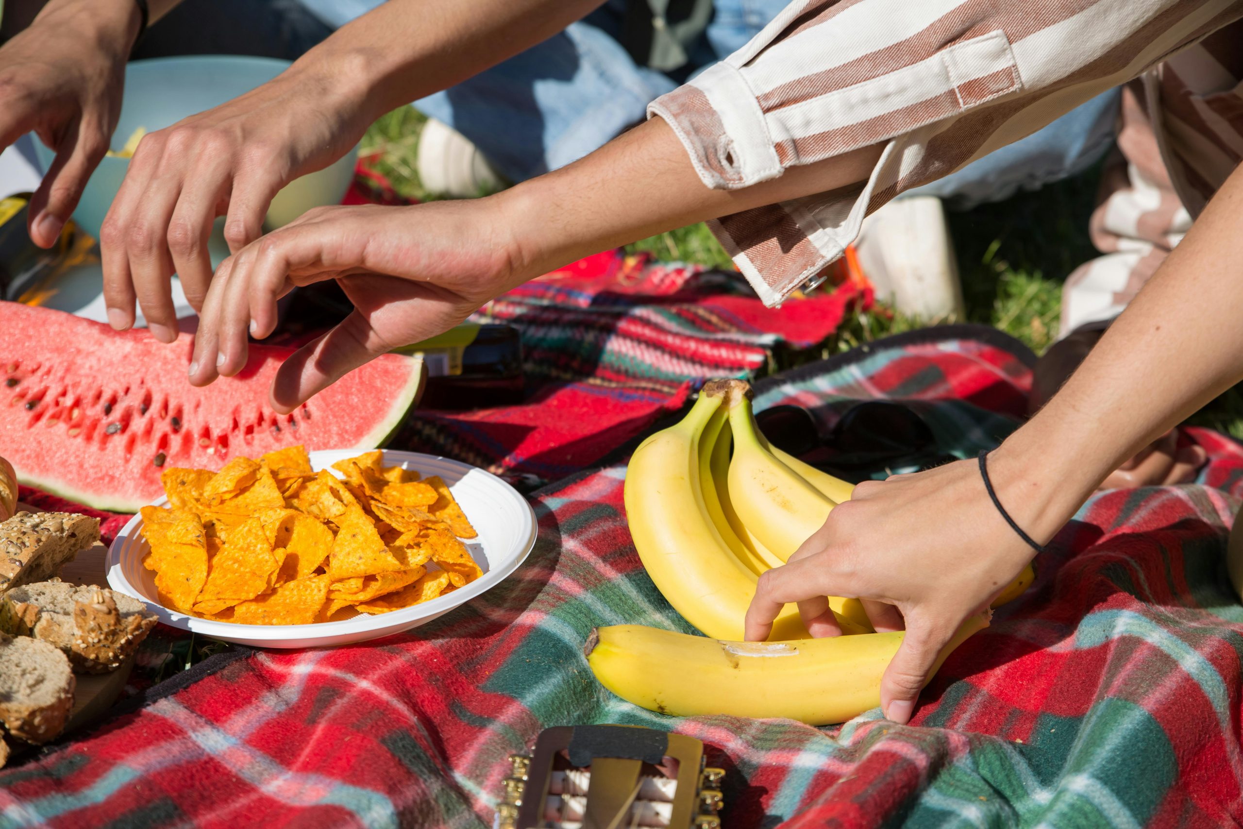 Hands reaching for snacks and fruits on a picnic blanket during a sunny day in Portugal.