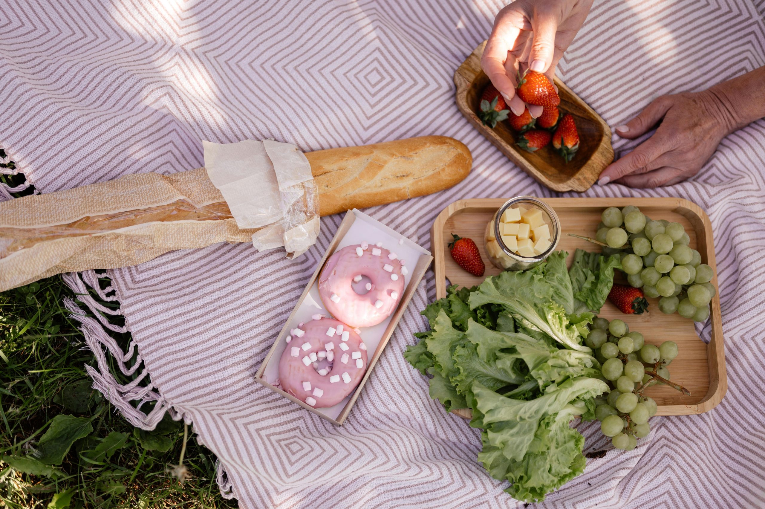 A summer picnic with baguette, pink donuts, grapes, and strawberries on a striped blanket.